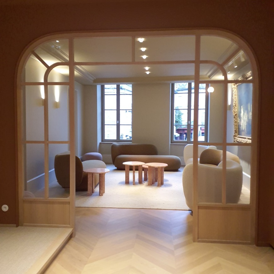 Salon vu à travers une verrière cintrée en bois clair, avec canapés arrondis et tables basses en pierre rose